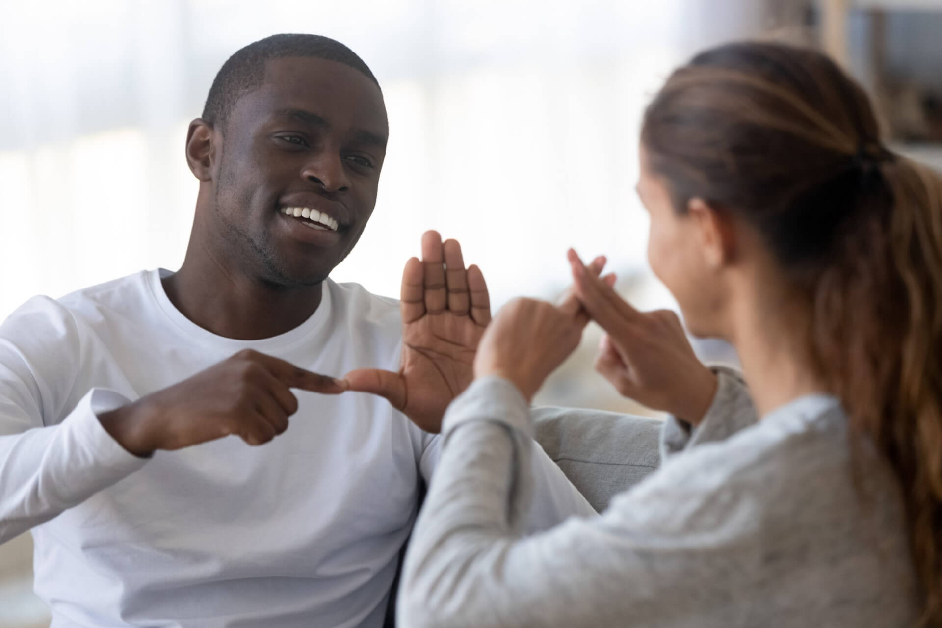 Smiling african American man sit on couch show hand gestures talking with female friend at home, international disabled hearing impaired couple or spouses use sign language communicating