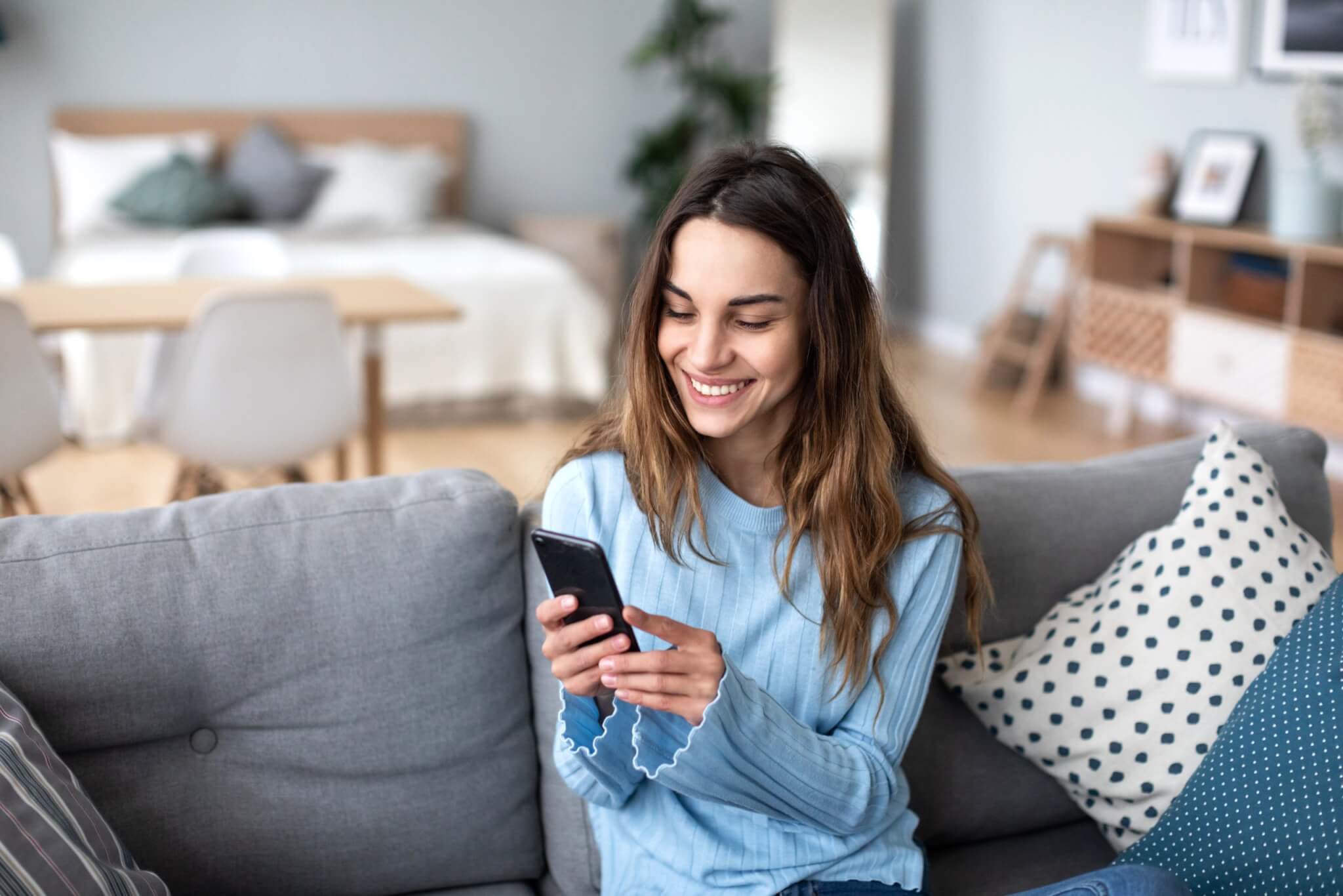 Cheerful young woman using mobile phone while sitting on a couch at home