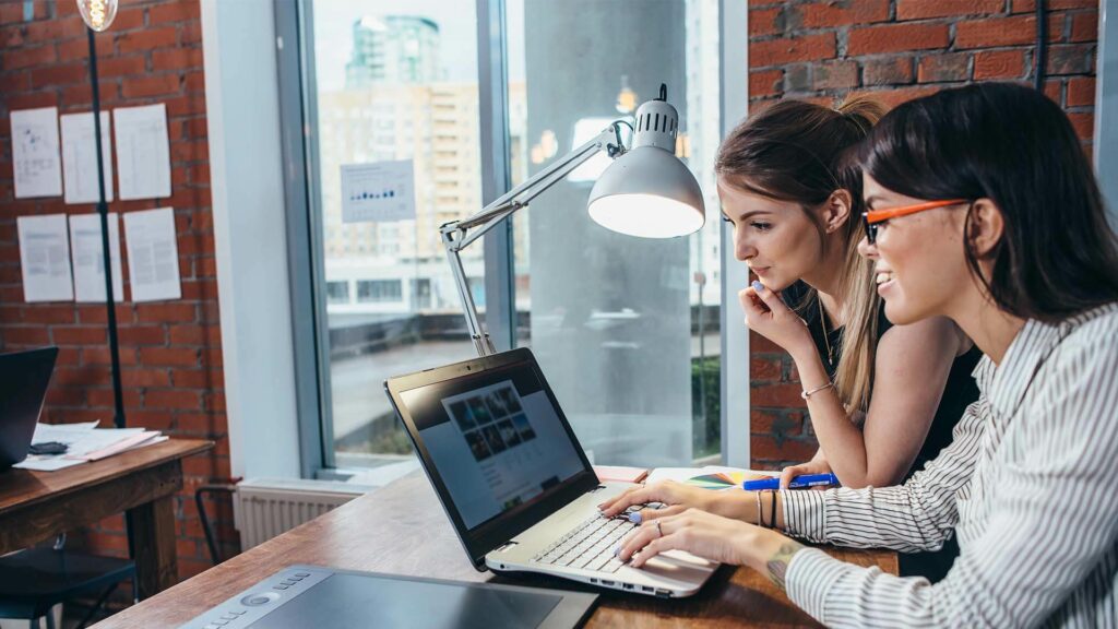 two woman looking at each other smiling and looking at laptop