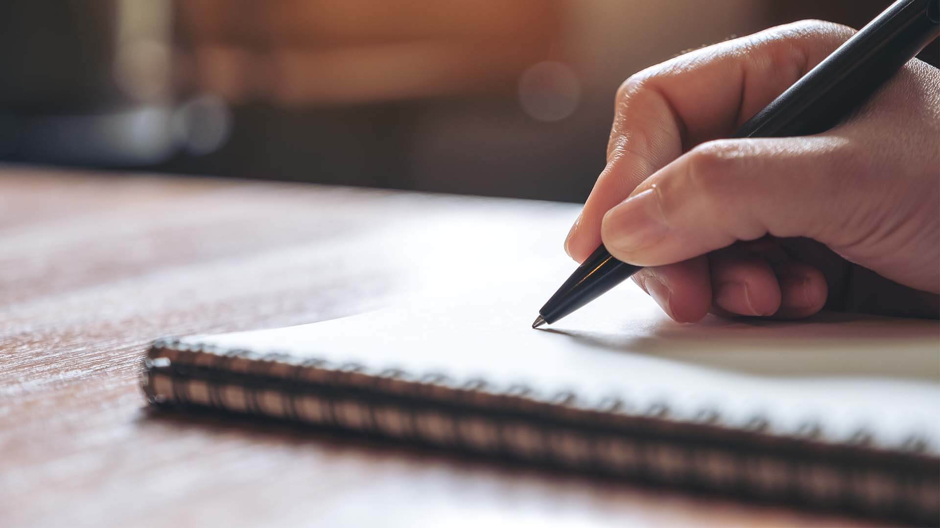 hand writing with a pen on a paper pad that is on a wooden table